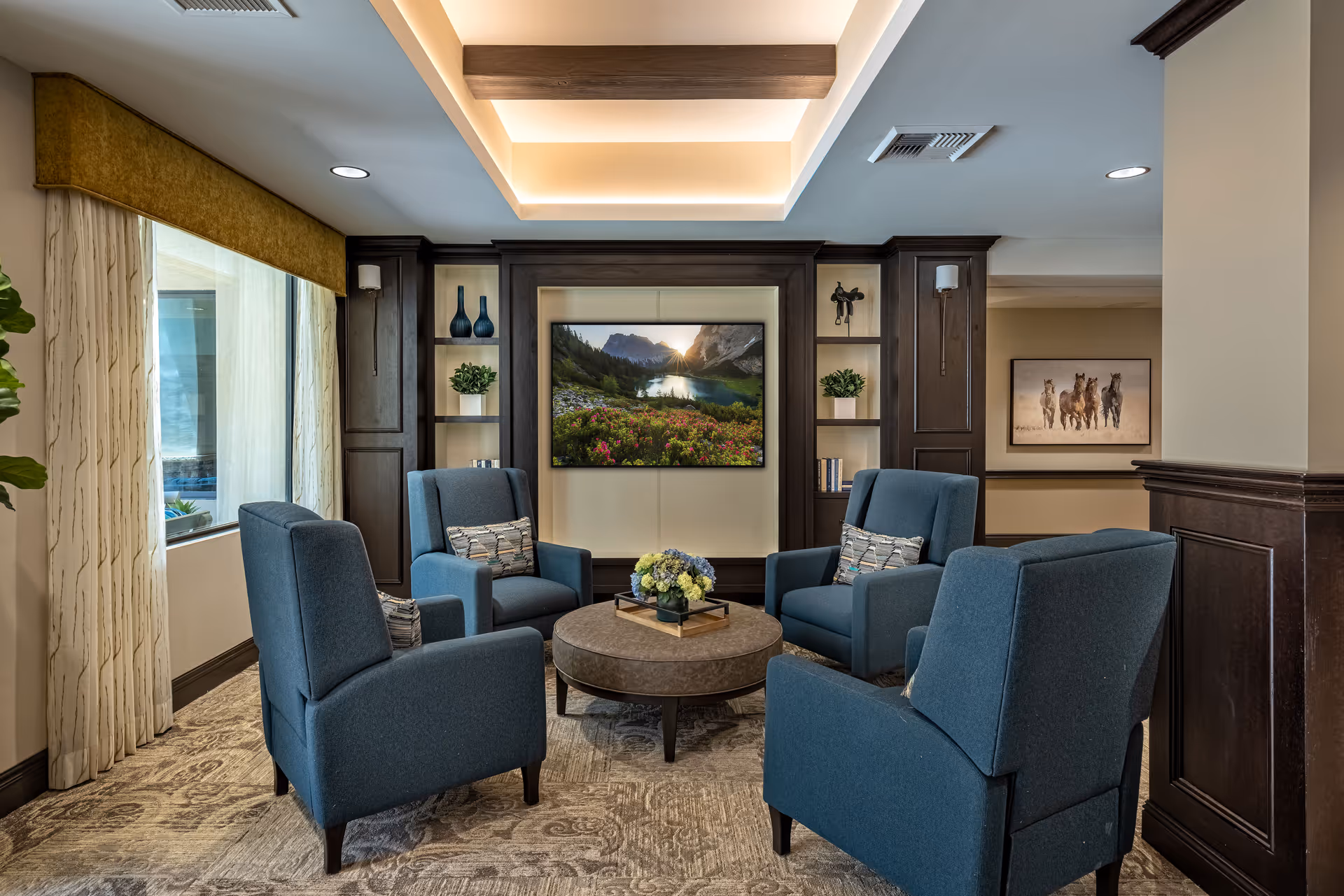 Comfortable seating area with four blue armchairs arranged around a round coffee table facing a wall-mounted TV in a well-lit lounge.