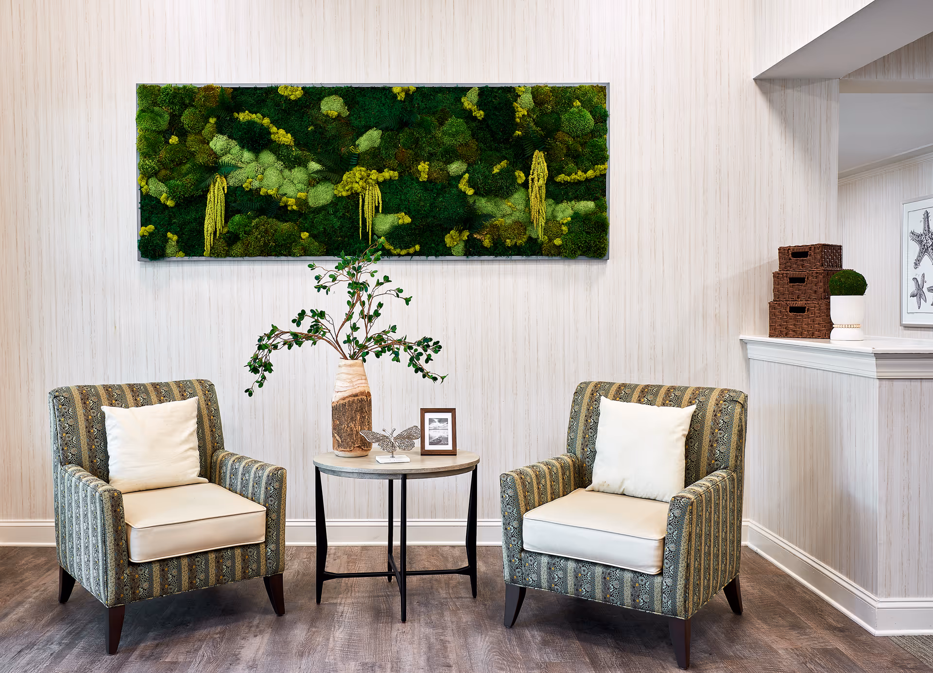 Two patterned armchairs with white cushions are placed on either side of a small round table with a vase of greenery, a butterfly ornament, and a small framed picture. Above the chairs is a rectangular wall art piece featuring green moss and plants. The room has light-colored walls and wood flooring.