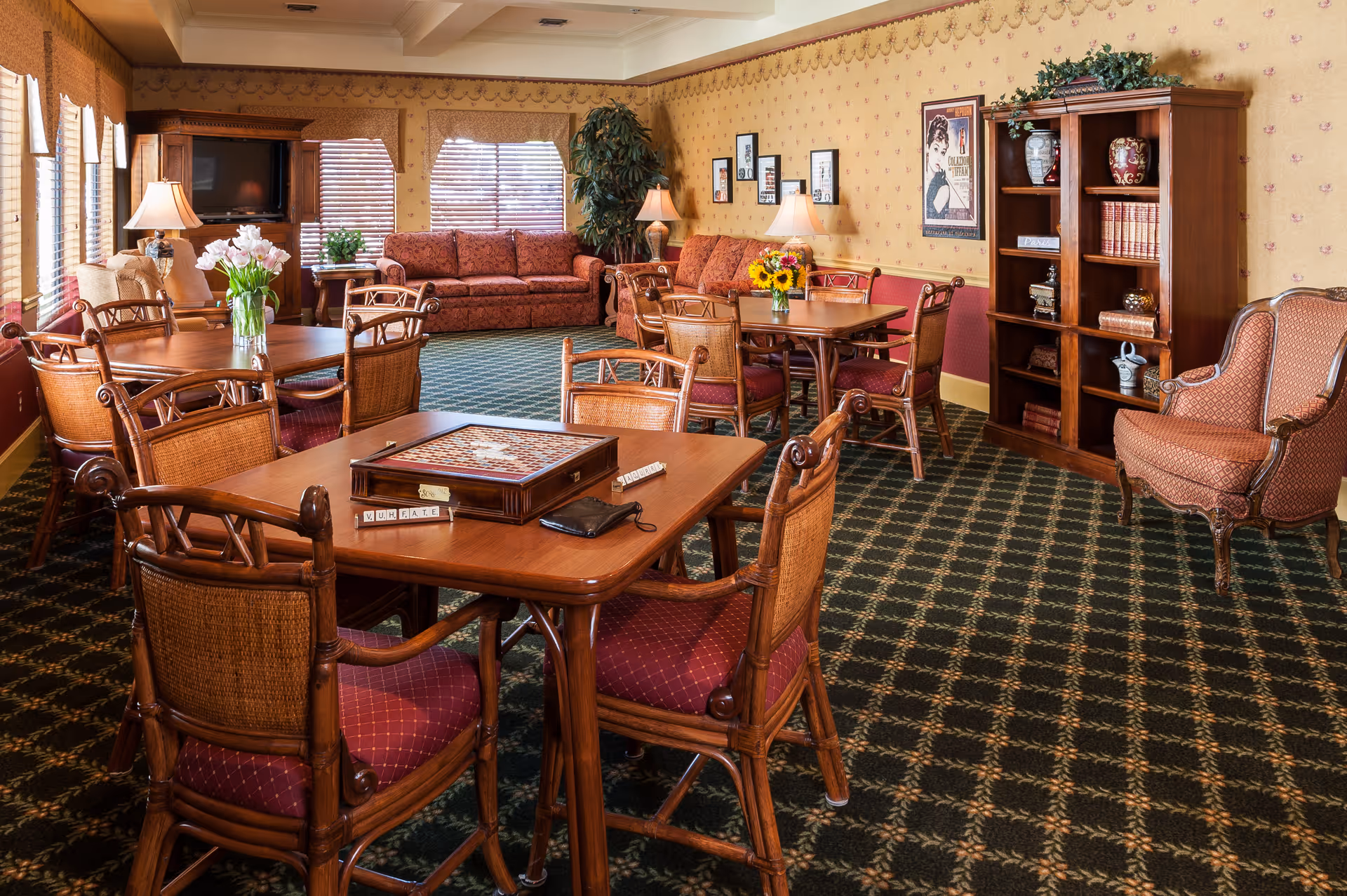 A cozy common area in a senior living facility with multiple wooden tables and chairs arranged for socializing or games. One table has a board game set up with Scrabble tiles. The room features patterned carpet, wallpaper with a floral border, framed pictures on the walls, a wooden bookshelf with decorative items, and two red upholstered sofas near windows with blinds. There are also table lamps and vases with flowers adding to the warm ambiance.