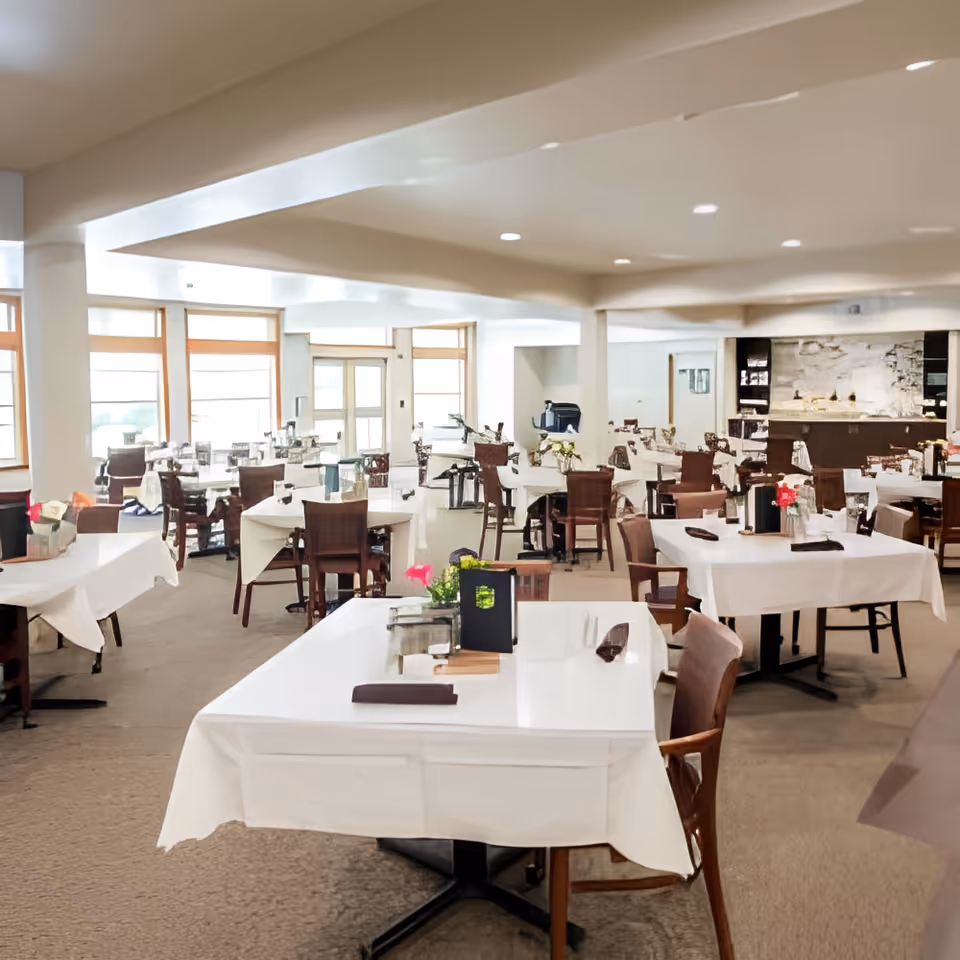 A spacious dining room with multiple tables covered in white tablecloths, each set with menus, napkins, and small flower arrangements. The room has large windows allowing natural light to fill the space, and wooden chairs are arranged around the tables. The background shows a counter area with shelves and a stone wall feature.