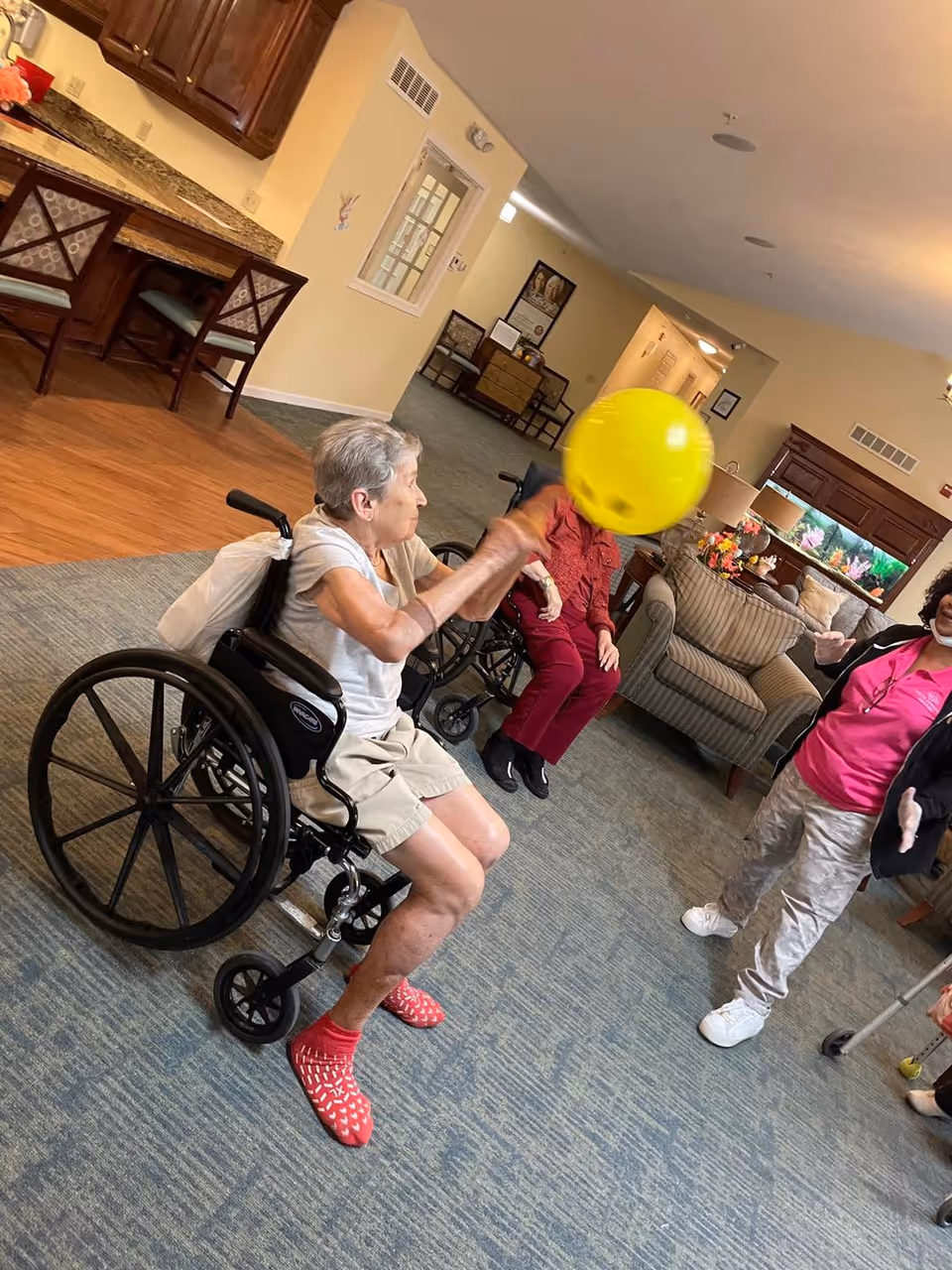 An elderly woman in a wheelchair plays with a yellow balloon in a senior living common room with other residents and staff.
