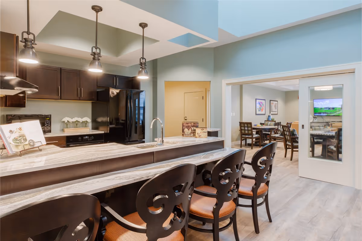 Modern kitchen area with a marble countertop island featuring four dark wooden chairs with cushioned seats. The kitchen has dark wood cabinets, a black refrigerator, a microwave, and three pendant lights hanging from the ceiling. In the background, there is a dining area with tables and chairs visible through a sliding glass door.