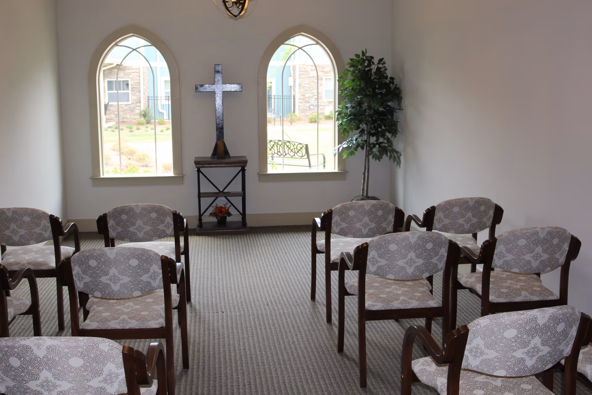 Small chapel room with patterned chairs arranged in rows facing a wooden cross on a stand between two arched windows. A potted plant is placed in the corner near the right window.