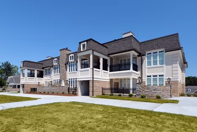 Exterior view of a modern two-story senior living facility building with balconies, large windows, stone and beige siding, and a well-maintained lawn under a clear blue sky.