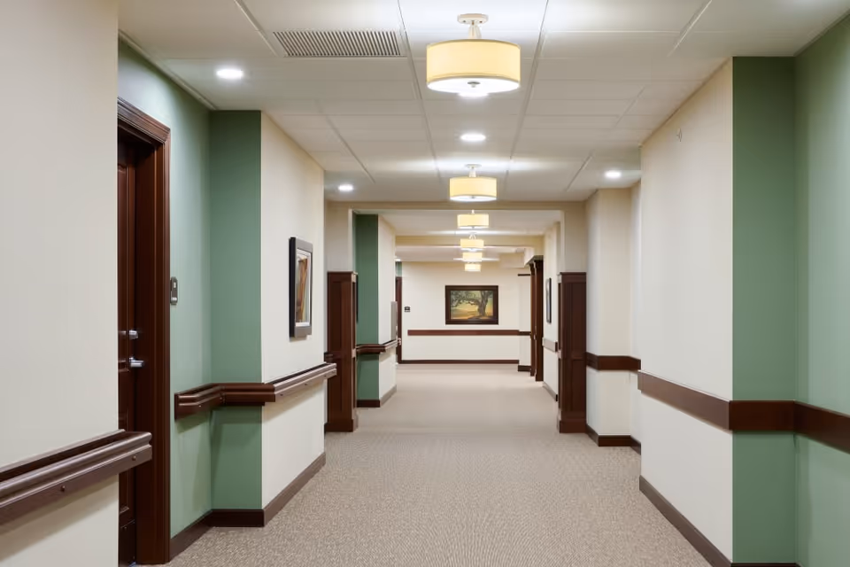 A well-lit hallway in a senior living facility with beige walls, green accent panels, and wooden handrails. The ceiling has recessed lighting and hanging light fixtures. There are framed paintings on the walls and several doorways along the corridor.