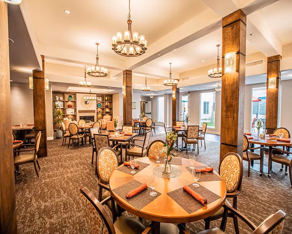 A bright and elegant dining room with round and square wooden tables set with placemats, folded orange napkins, and glassware. The room features patterned upholstered chairs, wooden pillars, multiple chandeliers, and large windows letting in natural light. There is a fireplace with decorative shelves in the background.