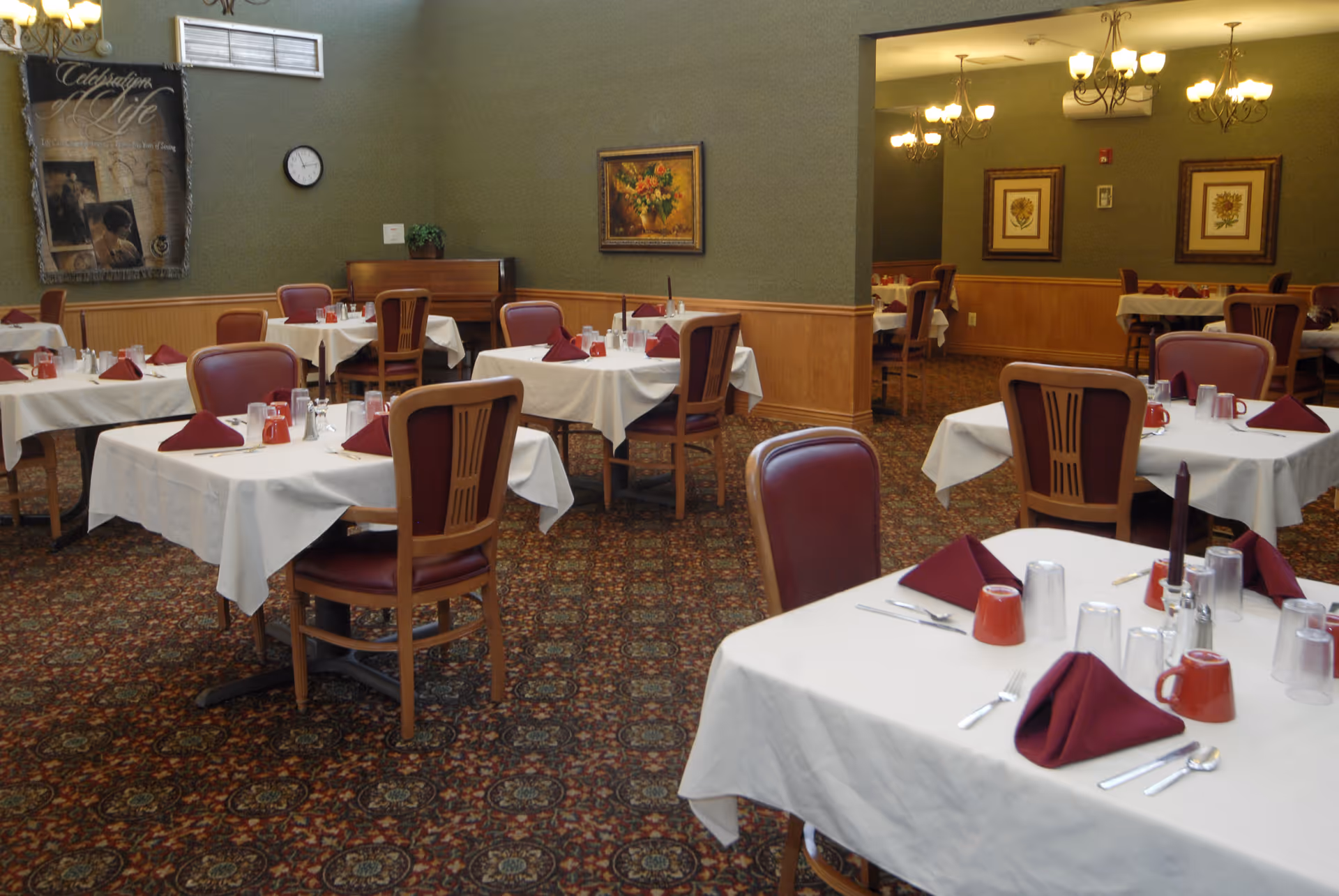A dining room with multiple tables covered in white tablecloths, each set with maroon napkins, silverware, glasses, and red mugs. The room has patterned carpet flooring, green walls with wooden wainscoting, framed artwork, and chandeliers providing warm lighting.