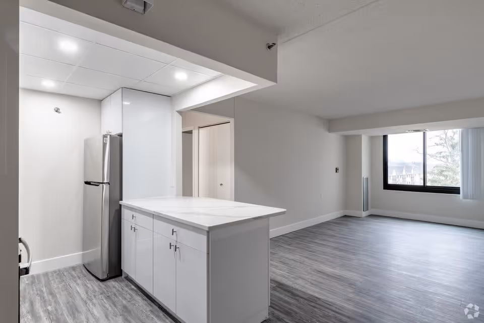 A modern, empty apartment interior featuring a kitchen area with white cabinets, a marble countertop island, and a stainless steel refrigerator. The kitchen opens into a spacious living area with large windows letting in natural light and gray wood-style flooring throughout.