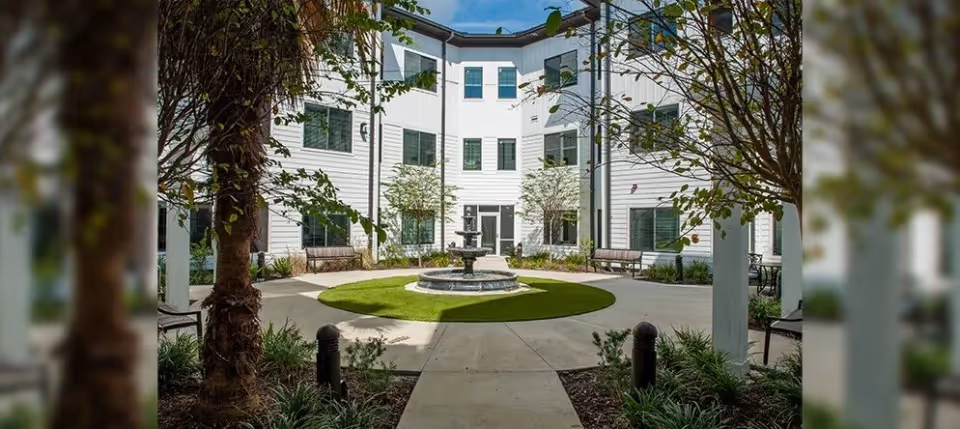 A three-story white building surrounding a landscaped courtyard with a central fountain, benches, and trees.