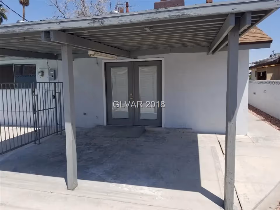 Covered outdoor patio area with a concrete floor and two supporting posts in front of a set of double glass doors. There is a metal gate to the left and a white wall with a small window. The sky is clear and blue.