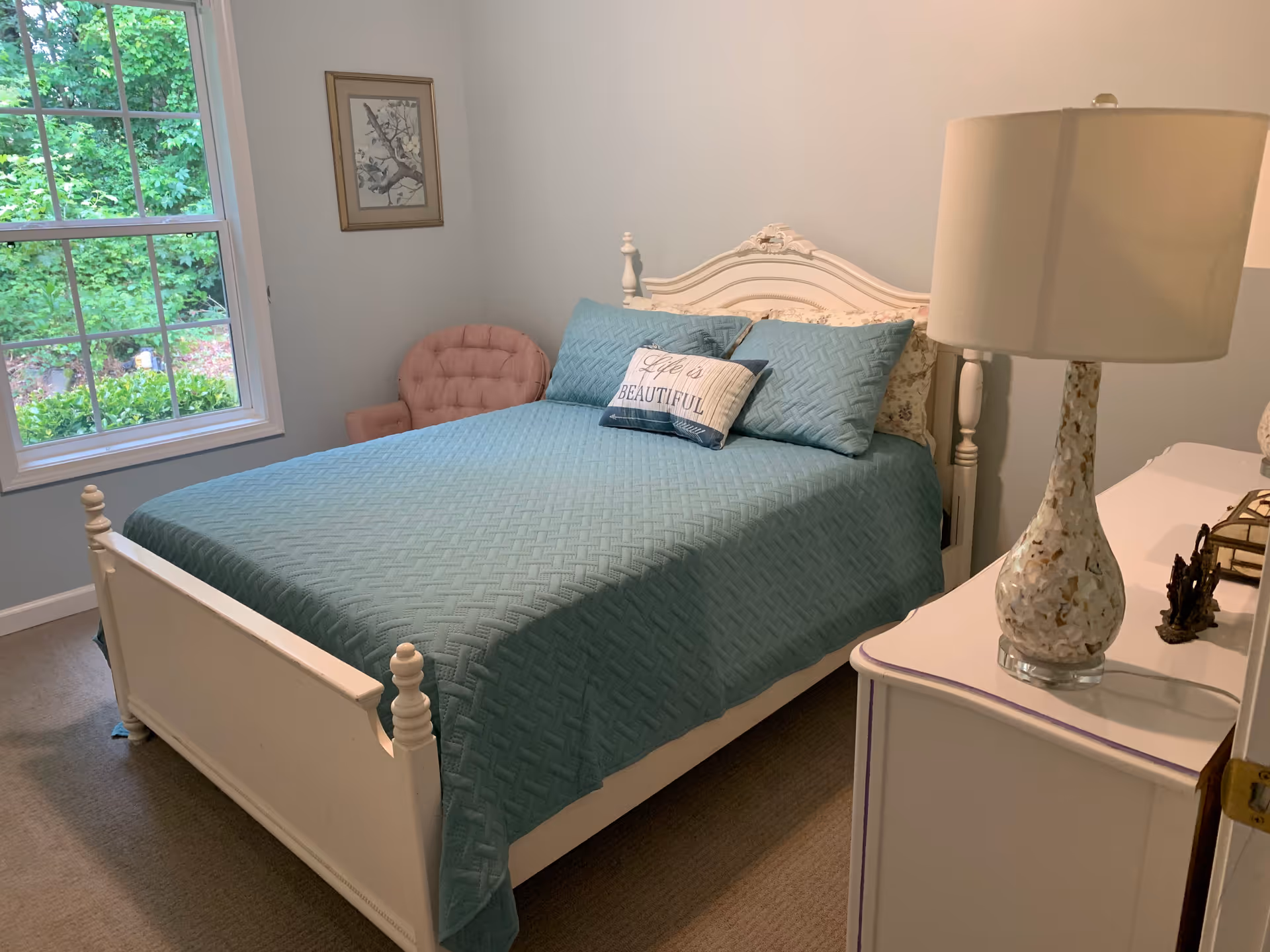 Cozy bedroom with a white wooden bed covered in a teal quilt, a decorative pillow, a nightstand with a lamp and a window showing greenery outside.