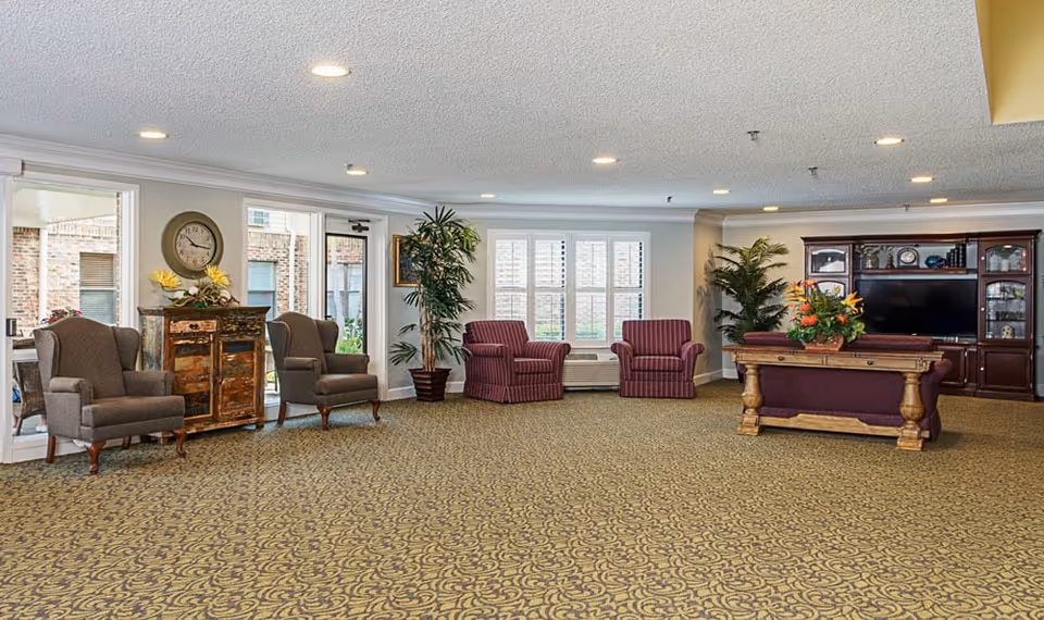 A spacious senior living facility common area with patterned carpet, several armchairs including two gray and two striped purple ones, a wooden cabinet with a clock and flowers on top, large windows with white shutters, potted plants, and a wooden entertainment center with a TV and decorative items.
