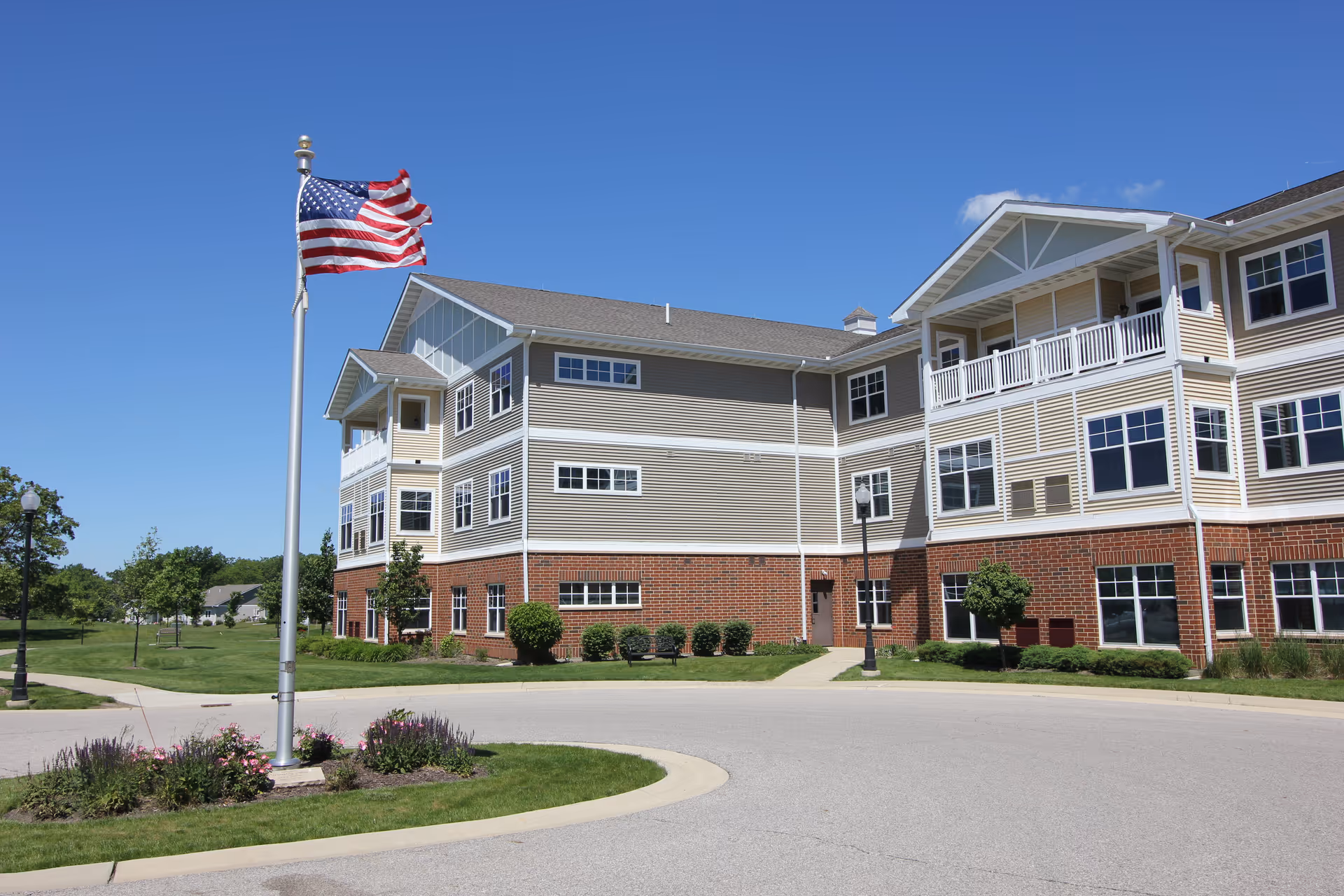 Exterior view of a multi-story senior living facility building with beige siding and red brick lower walls. An American flag flies on a flagpole in front of the building, surrounded by landscaped greenery and a curved driveway under a clear blue sky.