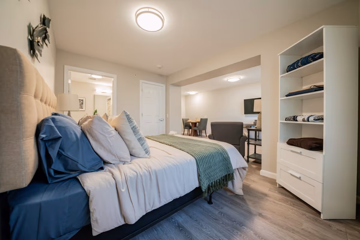 A cozy bedroom with a large bed featuring multiple pillows and a green throw blanket. To the right, there is a white shelving unit with folded blankets. In the background, an open doorway leads to a bathroom, and further back, there is a small dining area with a table and chairs, and a living area with a TV and armchair. The room has light-colored walls and wood flooring.