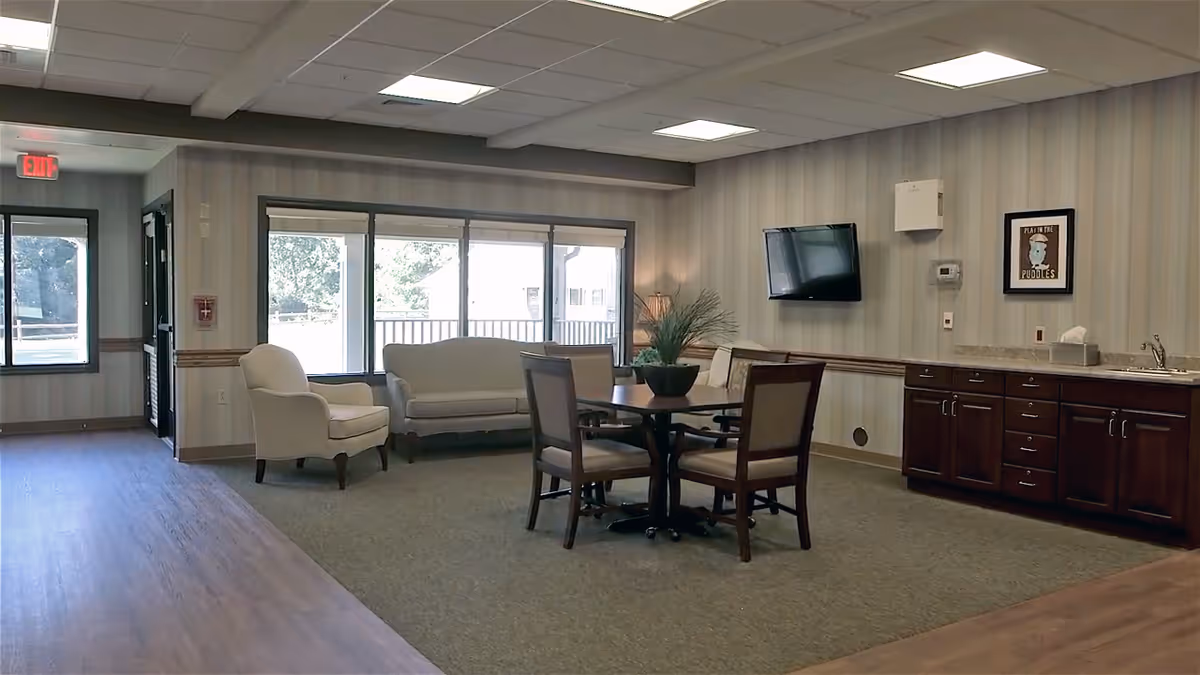 A common area in a senior living facility featuring a seating arrangement with a sofa, armchair, and a table with four chairs. The room has large windows letting in natural light, a wall-mounted TV, wooden cabinets with a countertop and sink, and a framed picture on the wall. The floor is a combination of carpet and wood, and there is an exit door visible in the background.