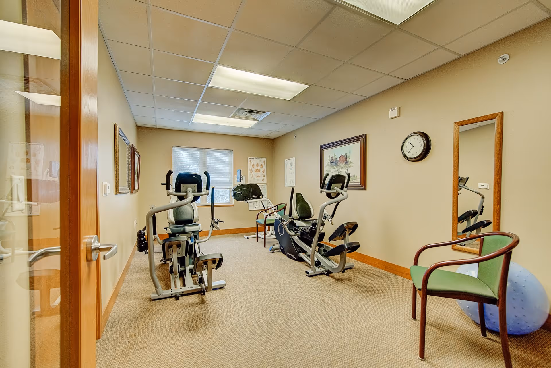 A small exercise room with two stationary recumbent bikes, a green chair, a large blue exercise ball, a wall clock, framed pictures, and a window letting in natural light.