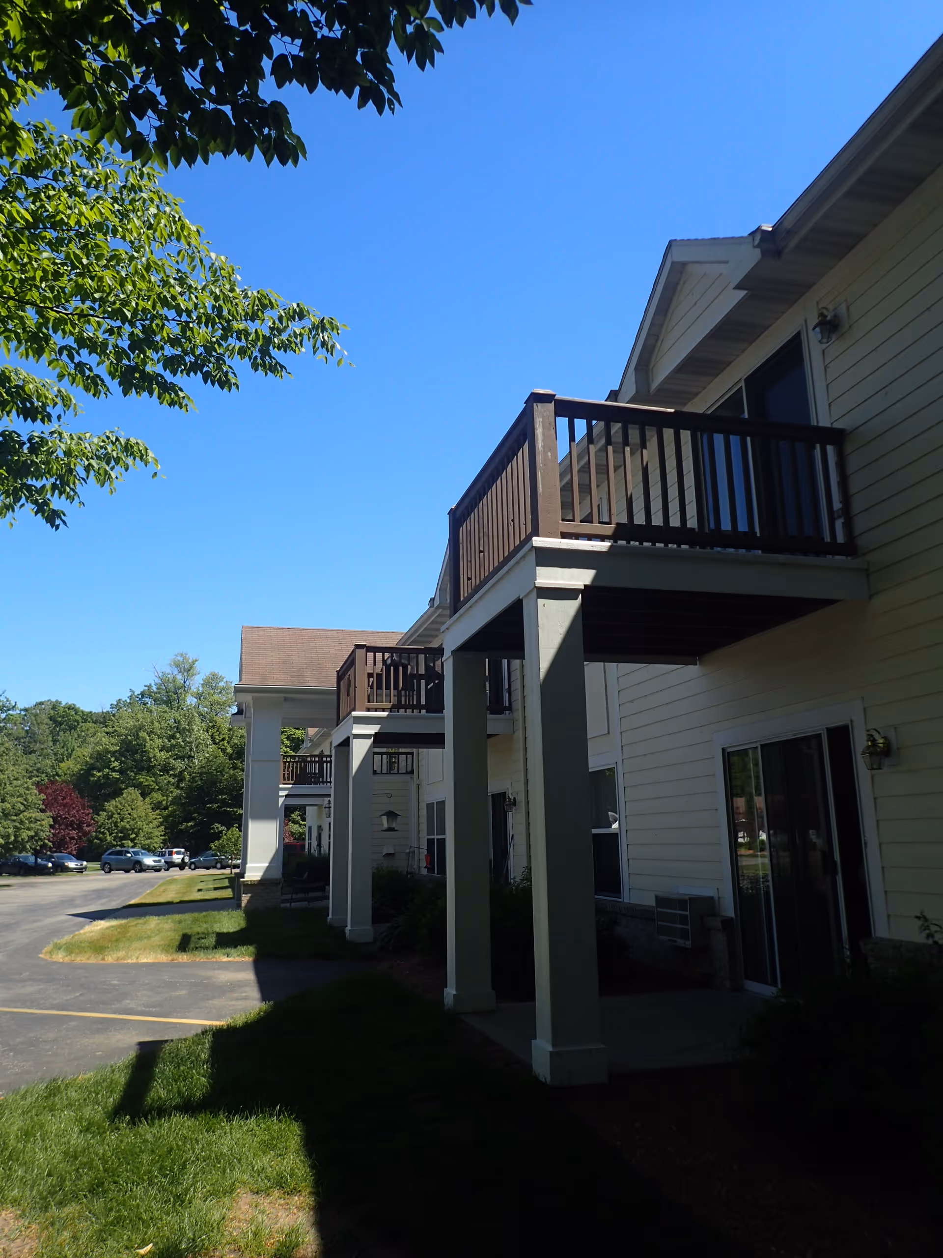 Exterior view of DaySpring Independent Living Apartments showing a row of two-story buildings with balconies on the upper floors and sliding glass doors on the ground floor. The buildings are light-colored with dark brown railings on the balconies. There is a paved driveway and green grass with trees under a clear blue sky.