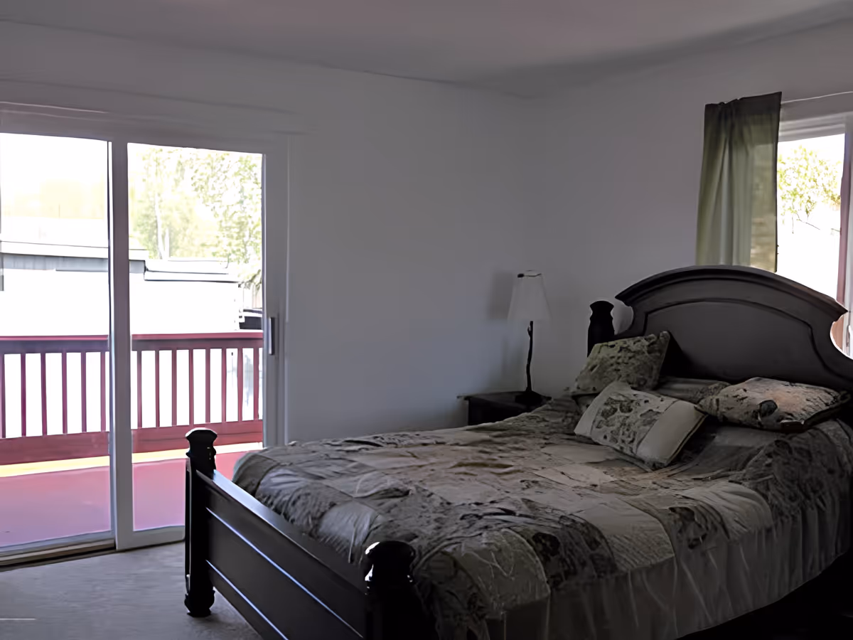 A bedroom with a dark wooden bed frame and a patterned quilt on the bed. There are multiple pillows on the bed. A small bedside table with a lamp is next to the bed. The room has a sliding glass door leading to a balcony with a red railing, and a window with a partially drawn curtain.