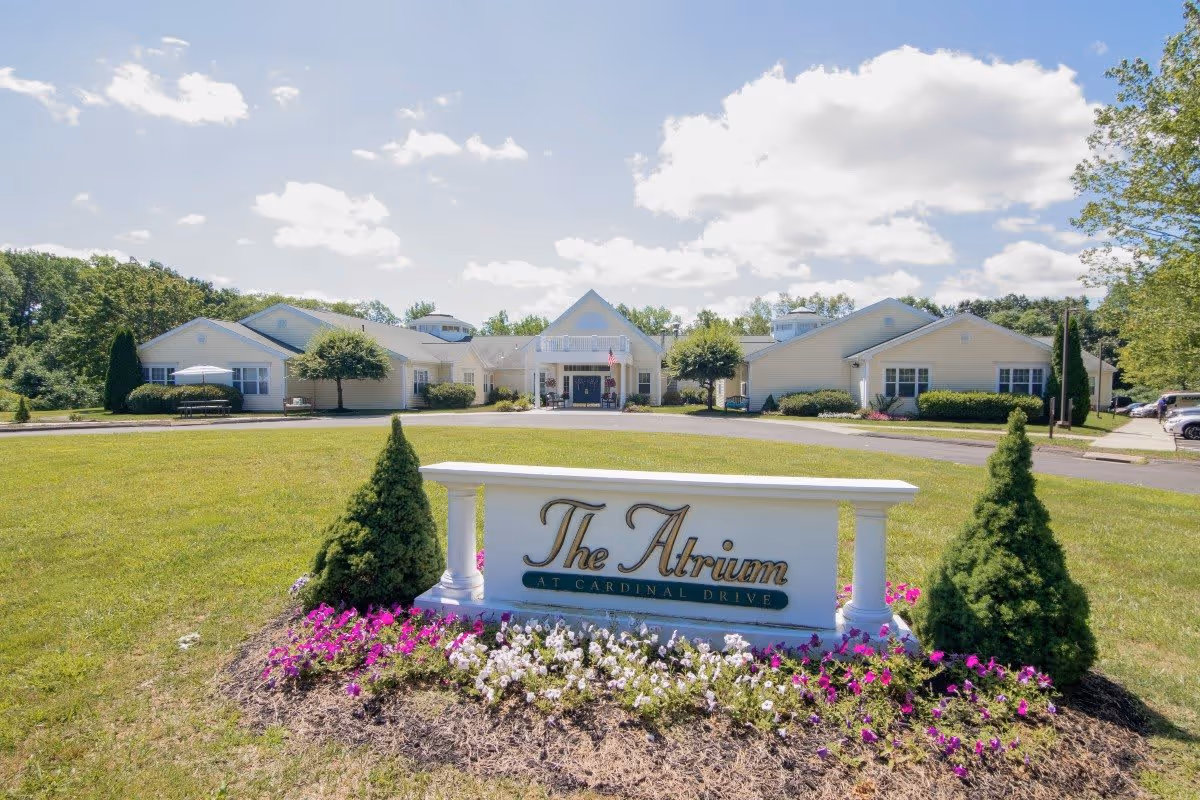 Exterior view of The Atrium at Cardinal Drive senior living facility with a large green lawn, flower beds, and a white sign displaying the facility's name in front. The building is light-colored with multiple sections and surrounded by trees under a partly cloudy sky.
