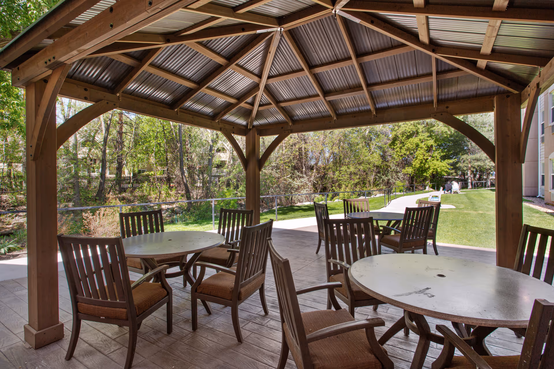 Outdoor covered patio area with round tables and wooden chairs, surrounded by greenery and trees, with a paved walkway and a building visible in the background.