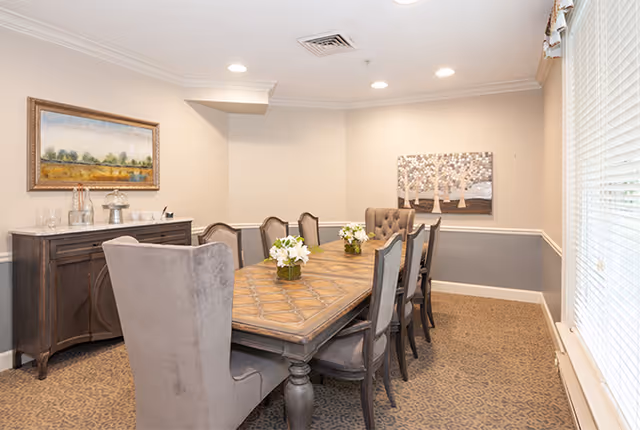 A well-lit dining room with a long wooden table surrounded by eight upholstered chairs. The table has three small flower arrangements as centerpieces. On the left side of the room, there is a dark wooden sideboard with glassware and decorative items. The walls are painted beige with a white chair rail, and two framed artworks hang on the walls. Large windows with blinds are on the right side, allowing natural light to fill the room.