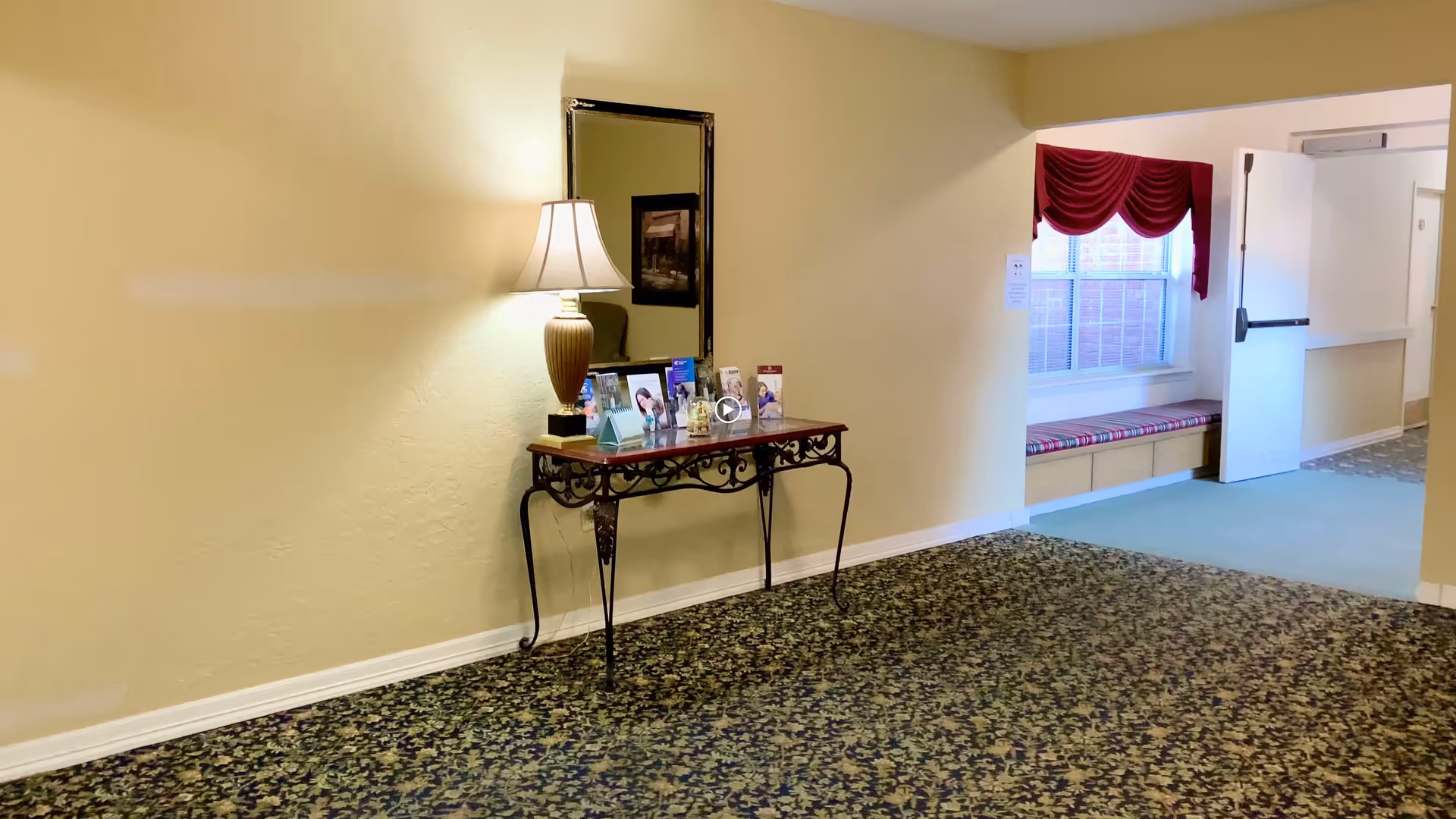 A hallway in a senior living facility with a decorative table against a yellow wall. On the table is a lamp, a mirror above it, and several framed photos and brochures. The hallway has patterned carpet and leads to a window seat with a red valance and a door that is open.