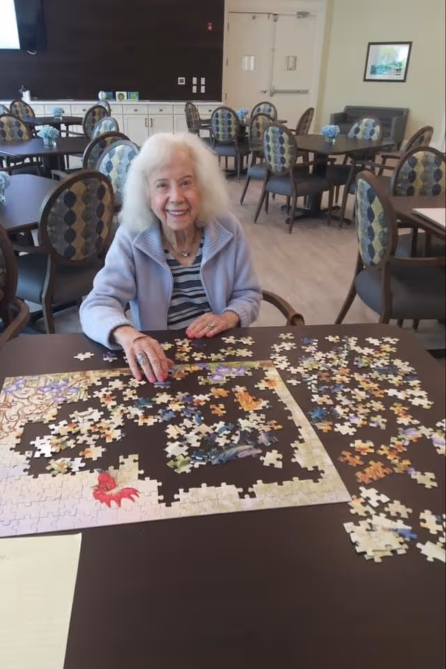An elderly woman smiling while assembling a jigsaw puzzle at a table in a communal dining/activity room.