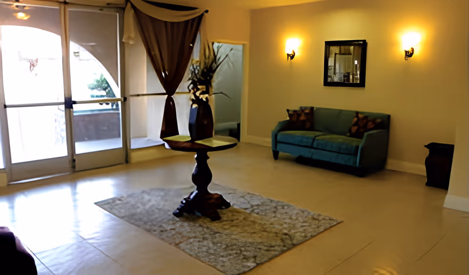 A well-lit assisted living facility lobby with a glass door entrance on the left, a small round wooden table with a vase of flowers in the center on a patterned rug, a teal upholstered sofa with two patterned cushions against the right wall, two wall sconces with warm lighting, and a mirror hanging between the sconces.