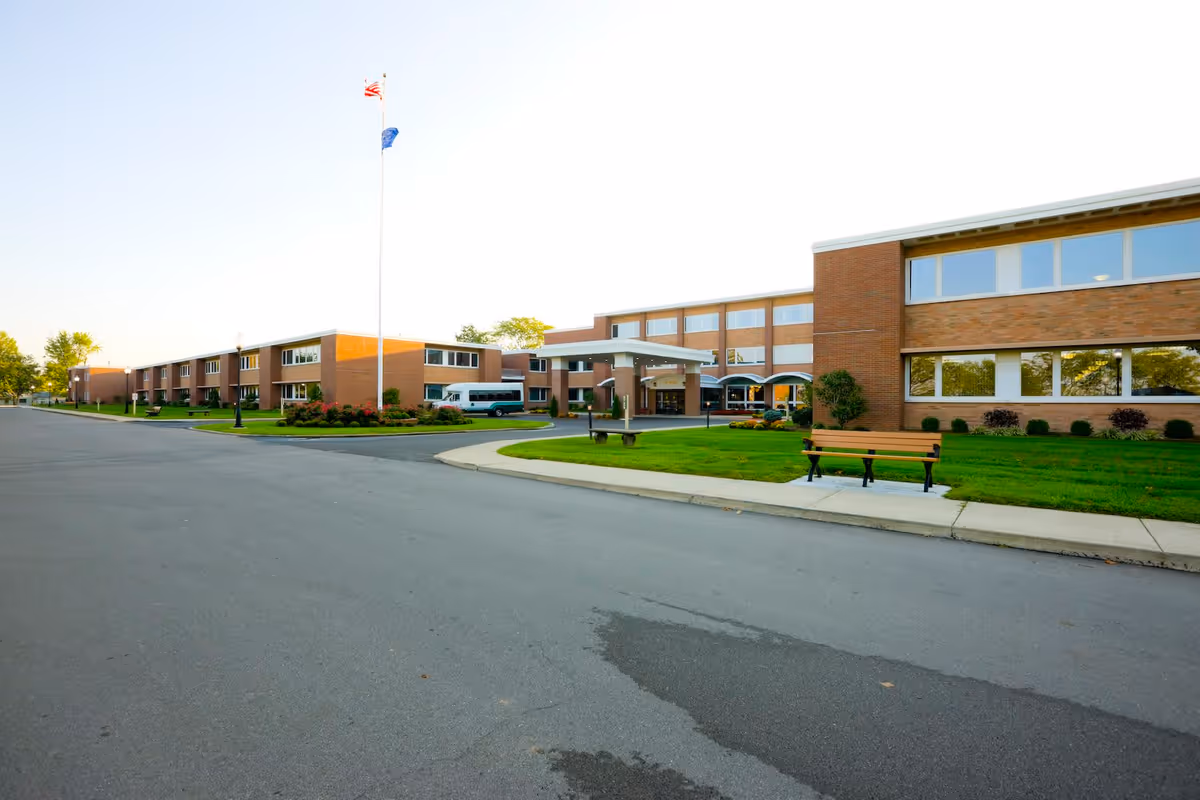 Exterior view of a large, two-story brick building with multiple windows, a covered entrance, a flagpole with two flags, a bench on a grassy area, and a paved driveway or road in front.