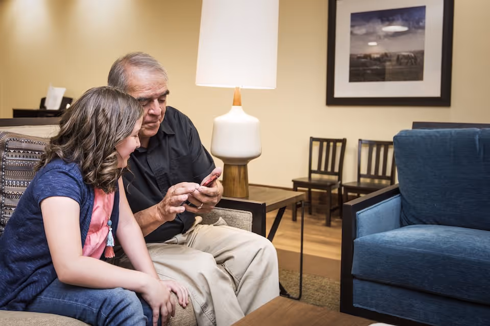 An elderly man and a young girl sitting together on a couch in a cozy living room. The man is showing something on a smartphone to the girl. The room has a table lamp on a side table, a framed picture on the wall, and several chairs in the background.