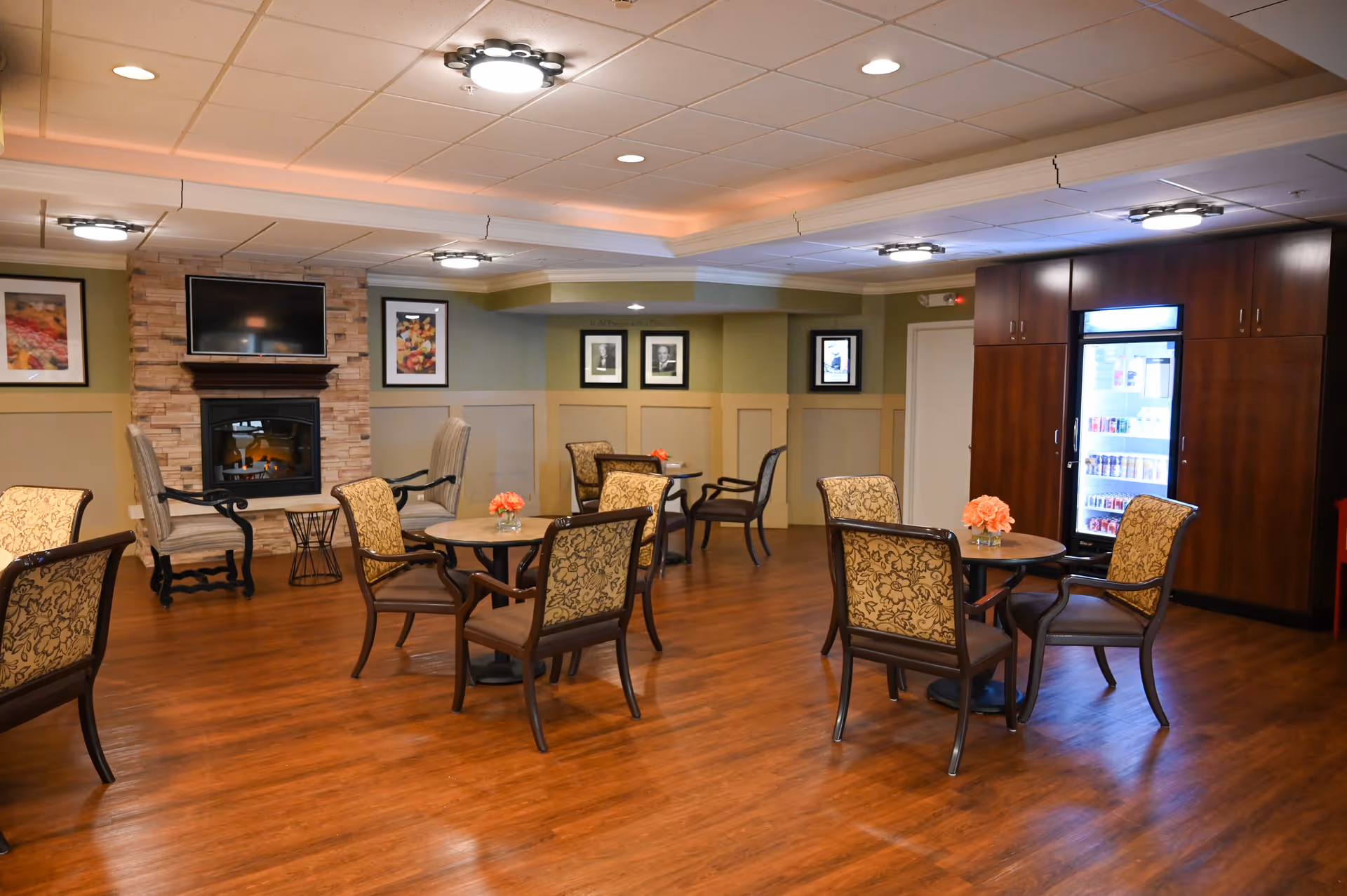 A cozy common area in a retirement community featuring several round tables with patterned chairs arranged around them. Each table has a small vase with orange flowers. The room has wood flooring, a stone fireplace with a mounted flat-screen TV above it, framed artwork on the walls, and a large wooden cabinet with a glass-front refrigerator stocked with beverages.