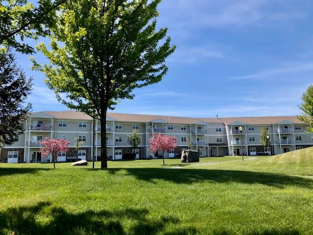 A three-story residential building with balconies, surrounded by a well-maintained green lawn with trees and flowering shrubs under a clear blue sky.