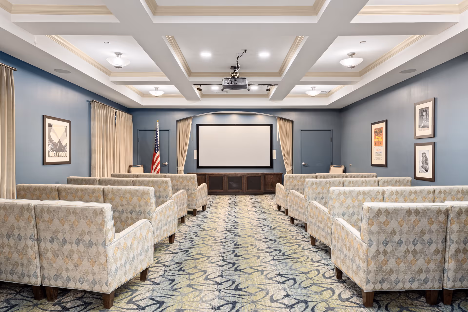 A small theater room with rows of patterned armchairs facing a large blank projection screen. The room has blue walls, framed pictures, beige curtains, and an American flag near the screen. The ceiling features recessed lighting and a projector mounted in the center.