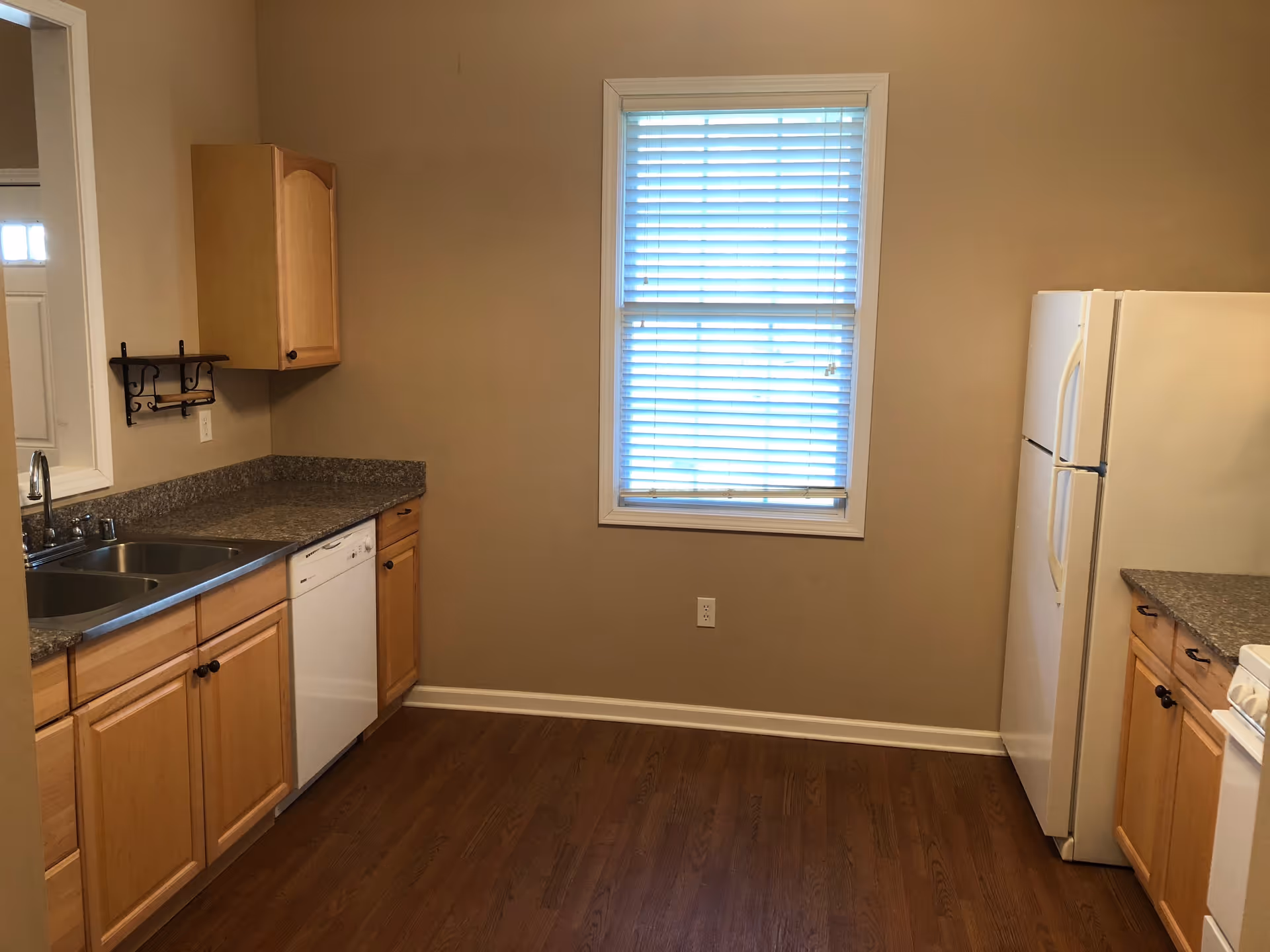 A kitchen with wooden cabinets, a double sink, a dishwasher, a white refrigerator, and a window with blinds. The floor is wooden and the walls are painted beige.