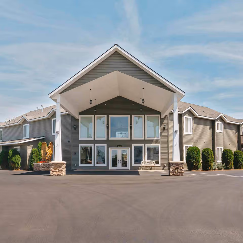 Front exterior of a two-story gray building with a large covered entrance, tall windows, columns, and landscaping.