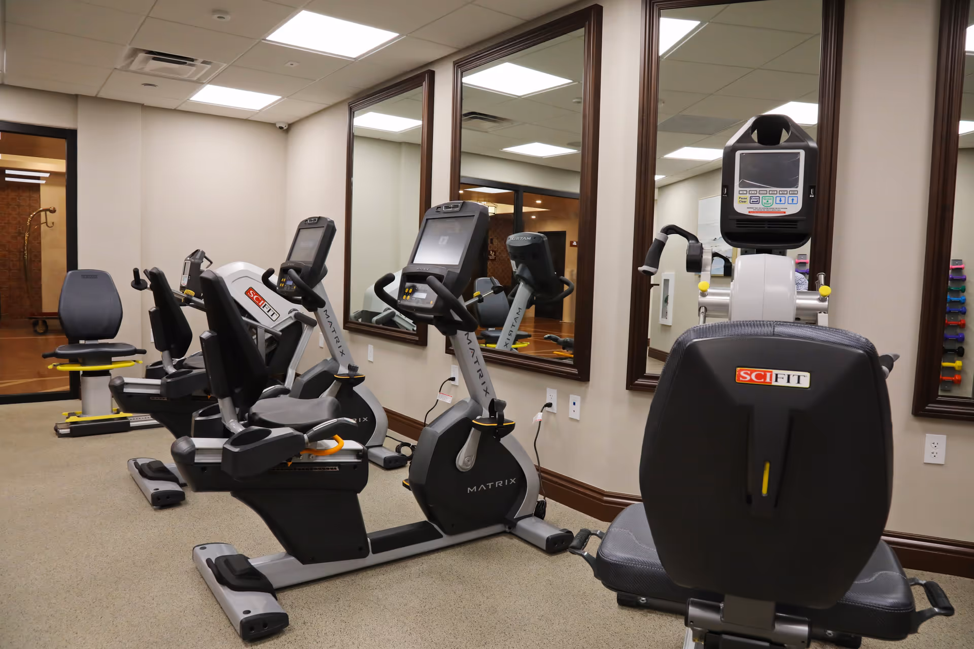 Indoor exercise room with stationary recumbent bikes and a large wall mirror reflecting the equipment and room. The room has beige walls, carpeted floor, and ceiling lights.
