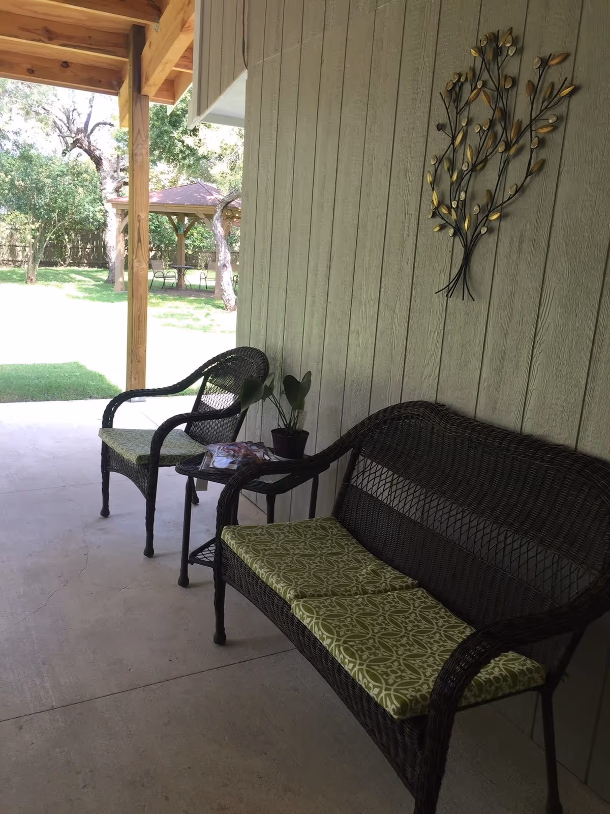 Covered outdoor patio area with two dark wicker chairs and a matching loveseat with green patterned cushions. A small table between the chairs holds a potted plant and some magazines. The wall behind the seating has a decorative metal wall art piece shaped like a tree with leaves. In the background, there is a grassy yard with trees and a gazebo with chairs and a table underneath.