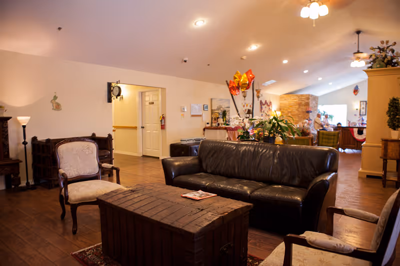 A cozy living room area in an assisted living facility featuring a black leather sofa, two upholstered armchairs, a wooden chest used as a coffee table, and various plants and decorations. The room has wooden flooring, soft lighting from ceiling fixtures, and a view into a hallway and another seating area in the background.