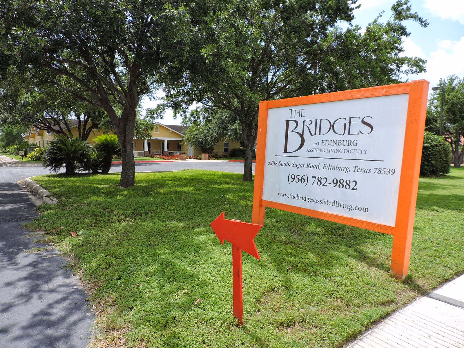Outdoor view of The Bridges at Edinburg Assisted Living Facility sign on a grassy area with trees and a driveway leading to a yellow building in the background. The sign includes the facility's name, address, phone number, and website, with a red arrow pointing left in front of it.