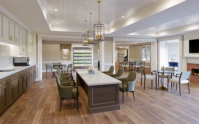 A spacious and well-lit common area with a large central table surrounded by green upholstered chairs, multiple smaller tables with chairs, wooden flooring, white walls, and a fireplace with a mounted TV above it. The room features modern pendant lighting and large windows allowing natural light to enter.