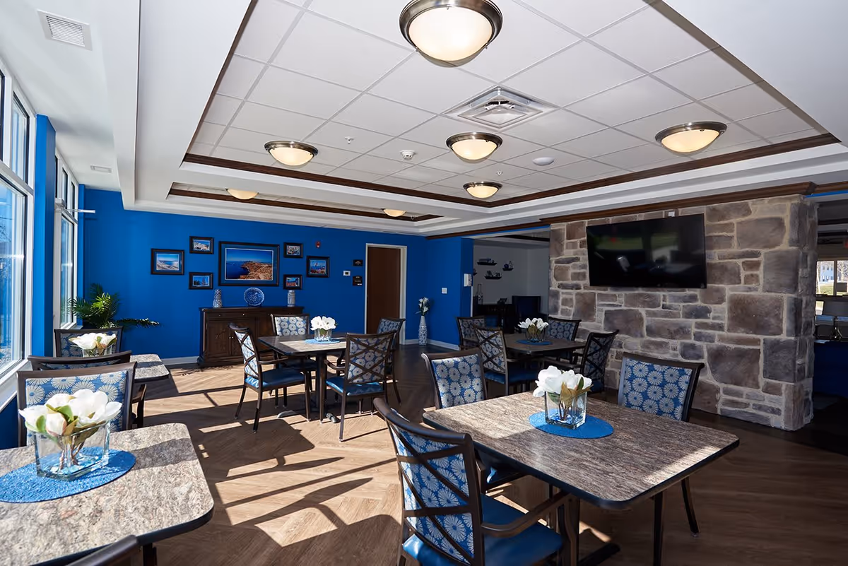 Bright communal dining area with tables and chairs, blue walls, a stone fireplace with mounted TV, and large windows.