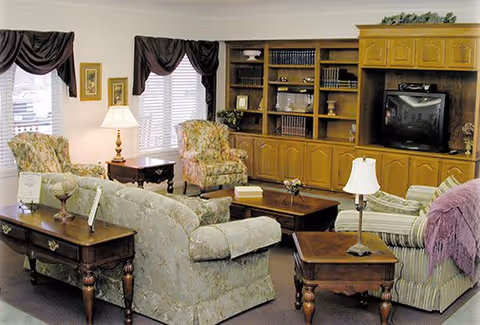 Sunlit communal living room with upholstered sofas and armchairs, wooden coffee and side tables, and a large built-in cabinet with a TV and bookshelves.
