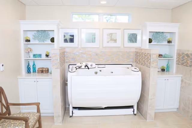 A bright and clean bathroom area featuring a white therapeutic bathtub centered between two half walls with mosaic tile accents. Behind the tub are built-in white shelves decorated with vases, plants, and framed artwork. A wooden chair with a patterned cushion is positioned to the left of the tub. The room has light-colored walls and floor tiles, with a small window above the artwork letting in natural light.