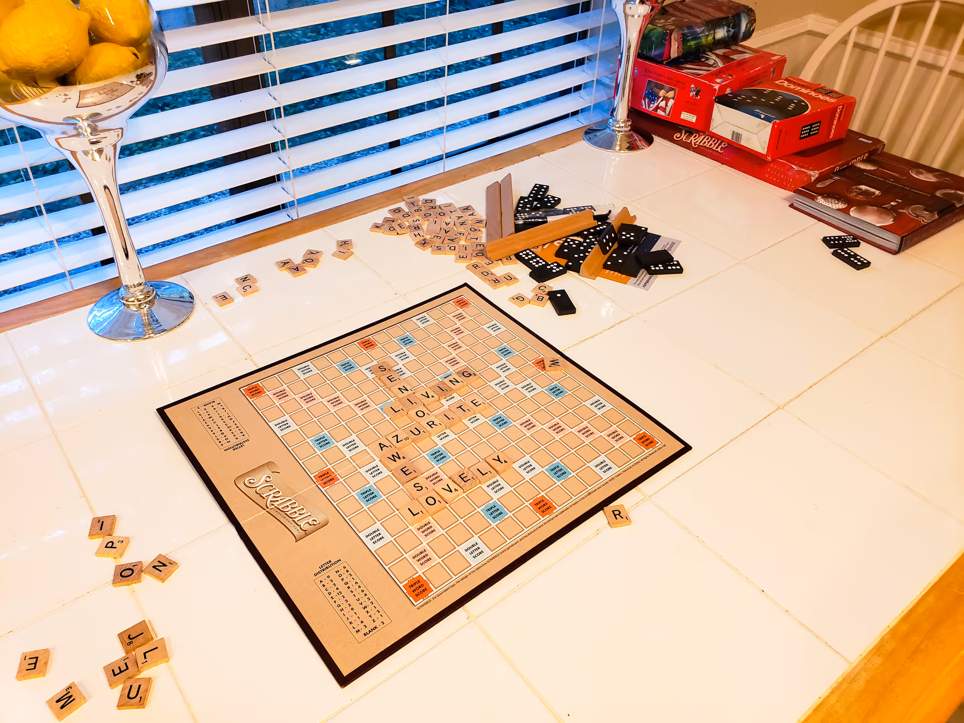 Scrabble board and scattered game pieces on a tiled table by window blinds, with dominoes, game boxes, and a fruit bowl nearby.