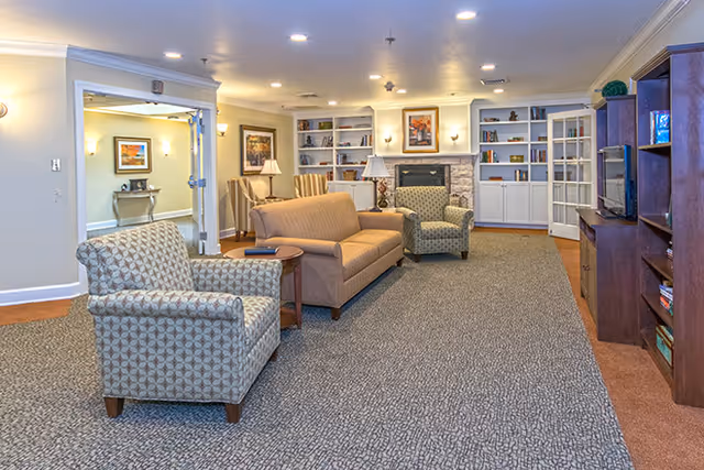 A cozy living room area in a senior living facility with patterned armchairs, a beige sofa, a wooden side table, built-in white bookshelves, a stone fireplace, framed artwork on the walls, and a wooden entertainment center with a TV.