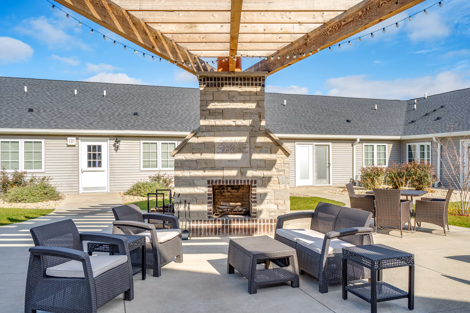 Outdoor patio area with a stone fireplace under a wooden pergola. The patio is furnished with dark wicker chairs and tables with light-colored cushions. The background shows a single-story building with gray siding, white doors, and windows, under a blue sky with some clouds.