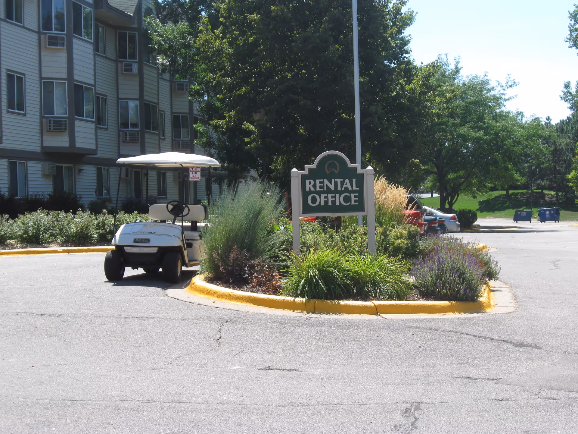 Outdoor view of a senior living facility parking area with a small white golf cart parked near a landscaped island. The island has a sign that reads 'RENTAL OFFICE' surrounded by green shrubs and plants. In the background, there are trees, parked cars, and part of a multi-story building with windows and air conditioning units.