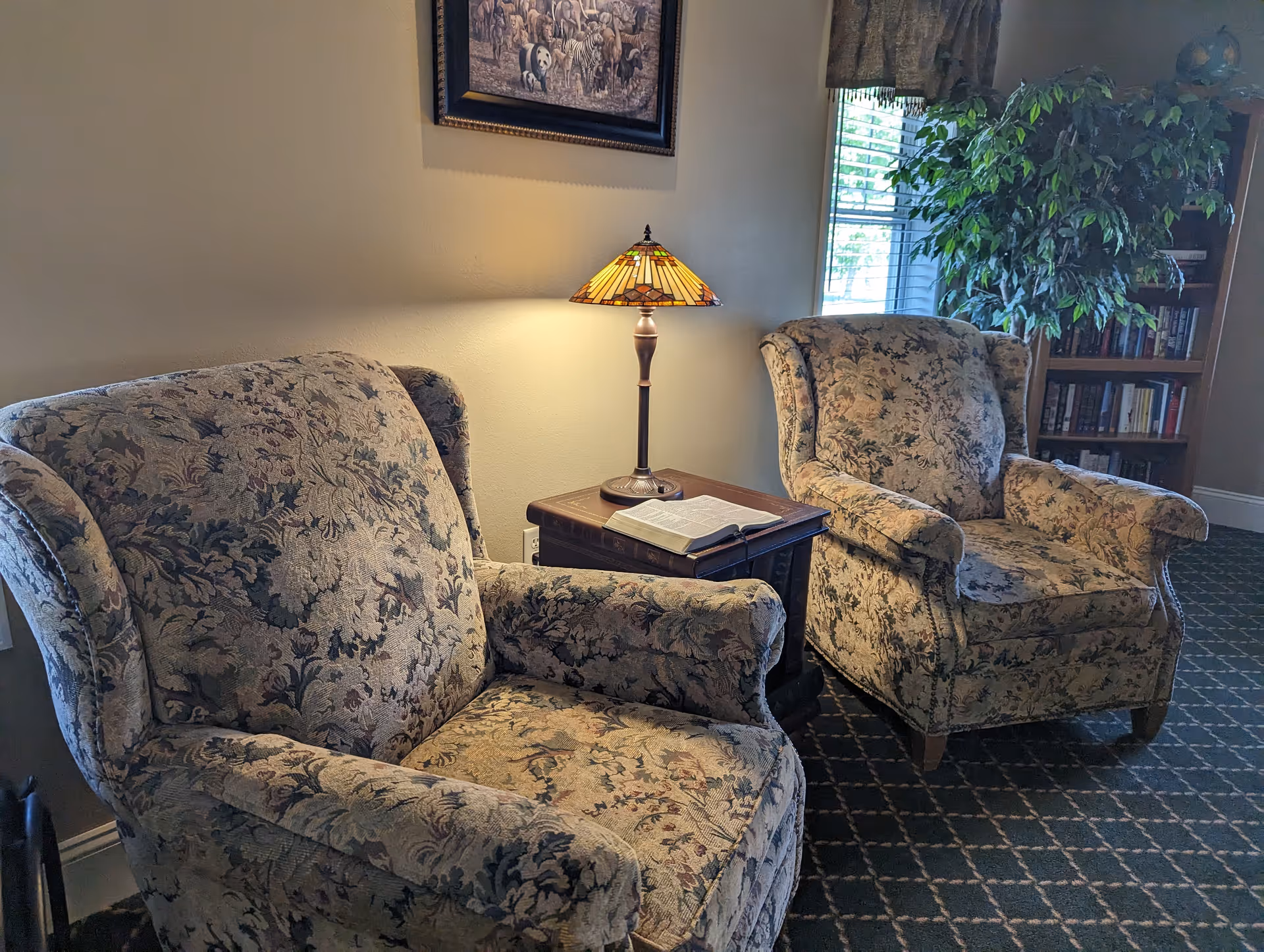 Two patterned upholstered armchairs with a wooden side table between them holding a Tiffany-style lamp and an open book, next to a window with blinds and a large leafy plant in a corner bookshelf filled with books.