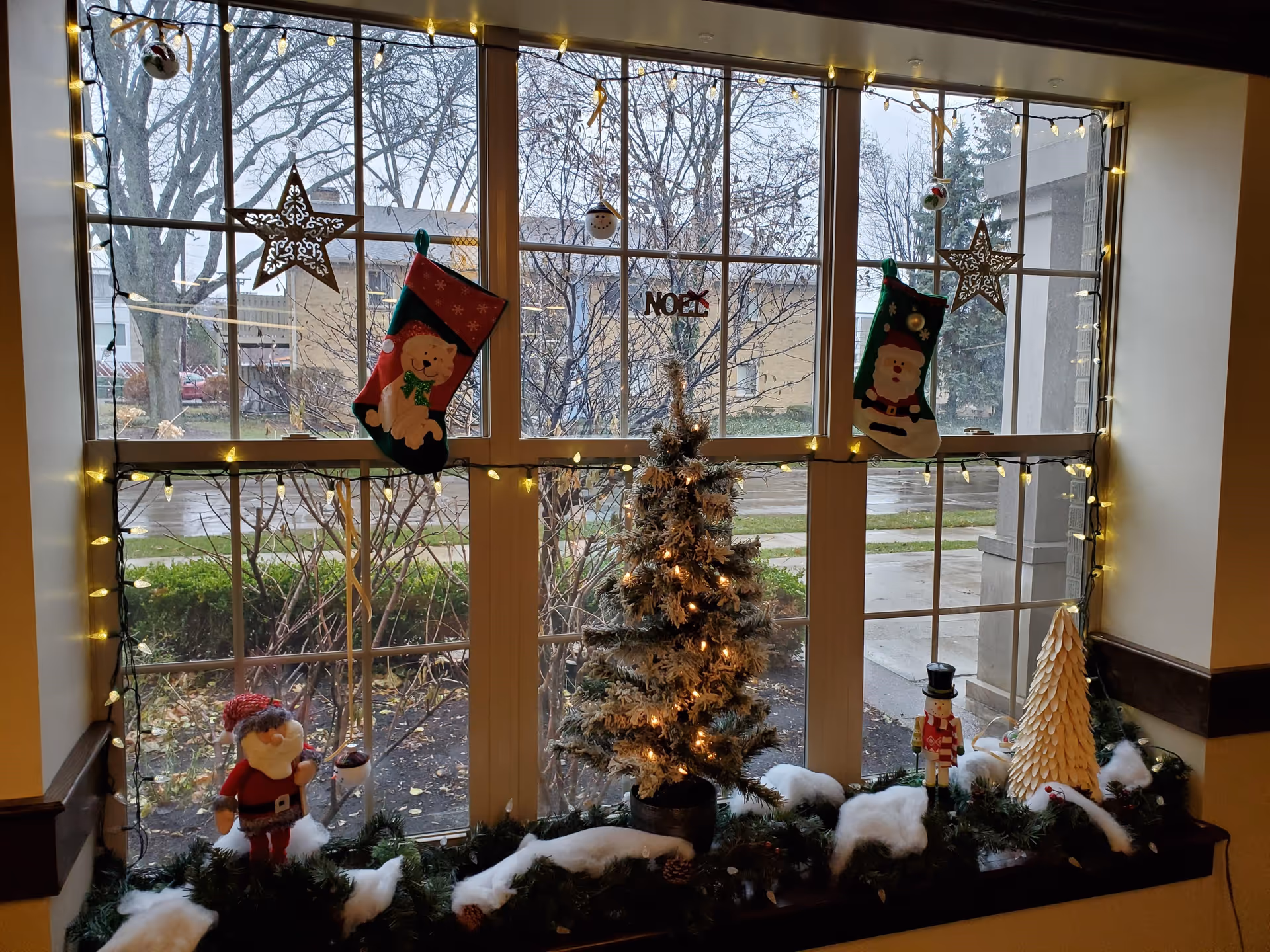 A large window decorated for Christmas with string lights, two hanging stockings, two star ornaments, a small decorated Christmas tree with lights, a Santa Claus figurine, a nutcracker figurine, and a white decorative tree. Outside the window, leafless trees and a building are visible on a cloudy day.