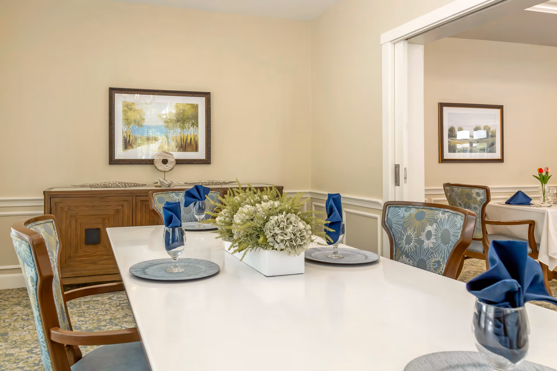 A bright formal dining room with a set white table, blue-napkin place settings, a central floral arrangement, and upholstered chairs.